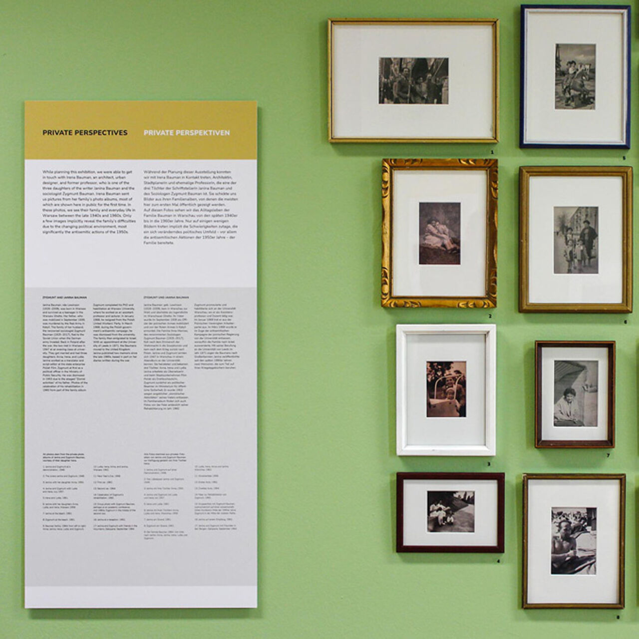 Mehrere schwarz-weiß Fotografien hängen vor einer grünen Wand. Sie sind alle in unterschiedlichen alten Rahmen gerahmt. Daneben hängt eine Informationstafel mit Text.