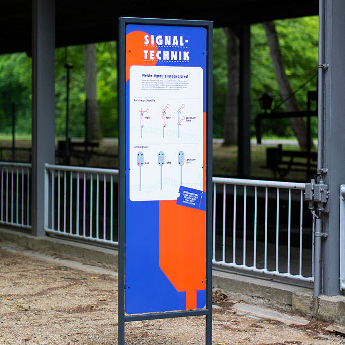 Ein Schild steht vor einem Bahnhof. Es ist orange-blau und gibt Auskunft über "Signaltechnik".