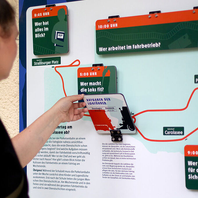 Zu sehen ist eine Informationstafel. Eine Hand klappt eine Klappe auf, unter der weiterführende Informationen zum Thema "Aufgaben der Lokführer" erscheinen. Darüber sind weitere dunkel grün und orangene Klappen zu erkennen, zu den Themen "Wer hat alles im Blick?", "Wer macht die Loks fit?" und "Wer arbeitet im Fahrbetrieb?".