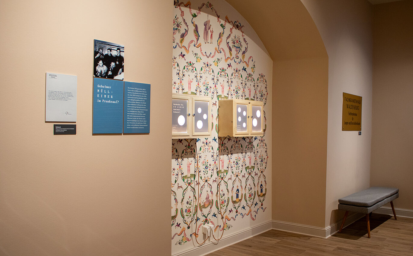 Präsentationsfläche In einer Ausbuchtung innerhalb einer Wand ist eine bunte florale und retro erscheinende Tapete zu erkennen. Vor dieser Tapete hängen zwei kleine beige Medizinschränke, welche aus Ausstellungsfläche verwendet werden. Links neben der Ausbuchtung an der Wand hängen unter einem schwarz-weiß Bild von fünf Männern zwei blaue Infotafeln, eine trägt die Überschrift "Geheimer Mülleimer im Prunksaal?". Rechts neben der Ausbuchtung ist unter einem goldenen Messing-Schild an der Wand eine graue Sitzbank.