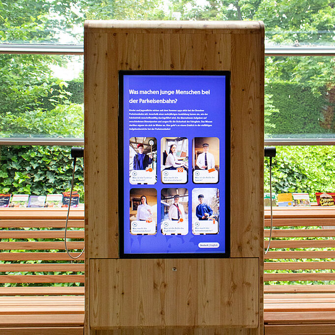 Eine Medienstation steht innerhalb des Bahnhofs der Dresdner Parkeisenbahn. Die Medienstation ist aus Holz und hat einen großen Bildschirm in der Mitte. An den Seiten sind Hörvorrichtungen zu erkennen. Der Bildschirm ist blau mit einigen Bildern darauf. Als Überschrift steht dort "Was machen junge Menschen bei der Parkeisenbahn?".