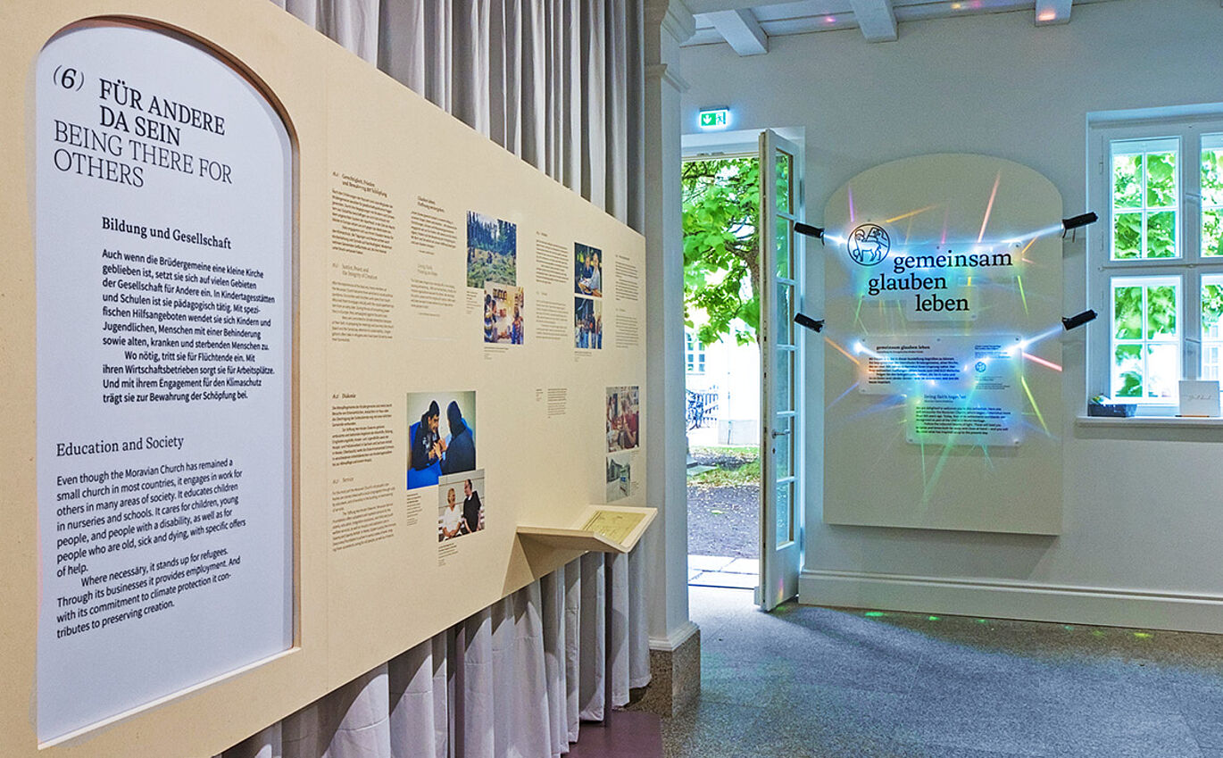 Foto der Ausstellung. Links ist eine Informationstafel zum Thema "Für andere da sein" zu erkennen. An der Wand daneben ist das angeleuchtete Key Visual zu sehen.