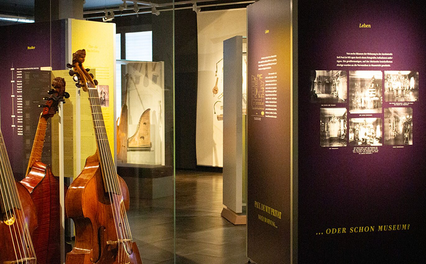 Auf der rechten Seite des Bildes ist eine lilane Informationstafel mit schwarz-weiß Fotografien zu erkennen. Links daneben in einem Schaukasten steht ein Musikinstrument.