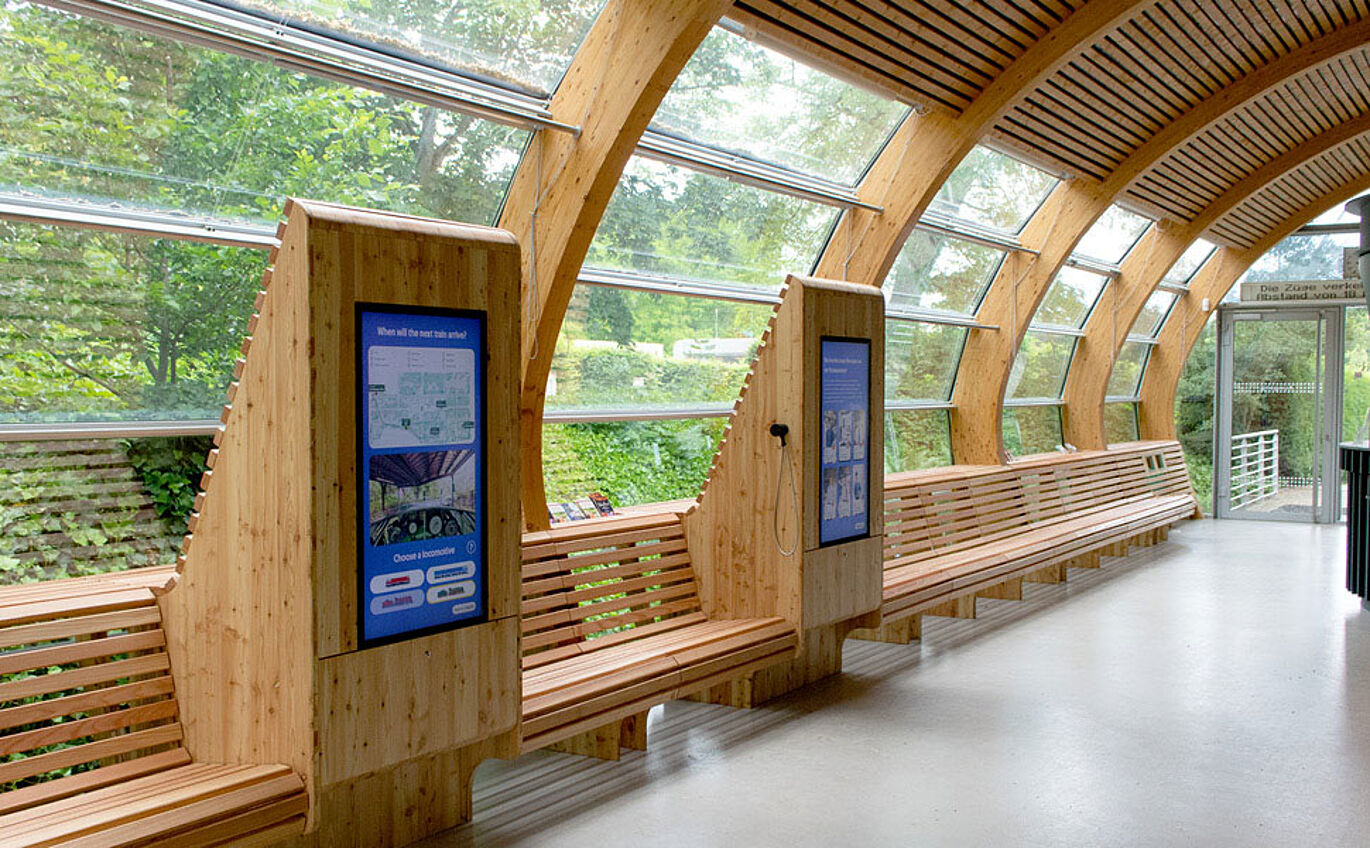 Der Bahnhof von innen ist zu sehen. Das Einrichtung erinnert an eine Wartehalle: Links stehen mehrere Bänke aus Holz vor einem großen Fenster. Zwischen den Bänken sind zwei Medienstationen - ebenfalls aus Holz - mit Bildschirmen zu erkennen. Die Decke ist ebenfalls aus Holz und spiegelt mit den langen Holzbalken das Aussehen der Bänke unten drunter wieder.