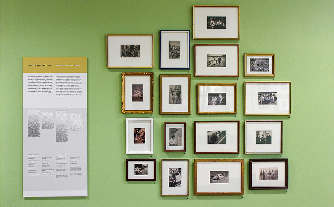 Mehrere schwarz-weiß Fotografien hängen an einer grünen Wand. Sie sind gerahmt, wobei jeder Rahmen unterschiedlich ist, viele der Rahmen sehen sehr alt aus. Links neben der Ansammlung an Fotos hängt eine Informationstafel.