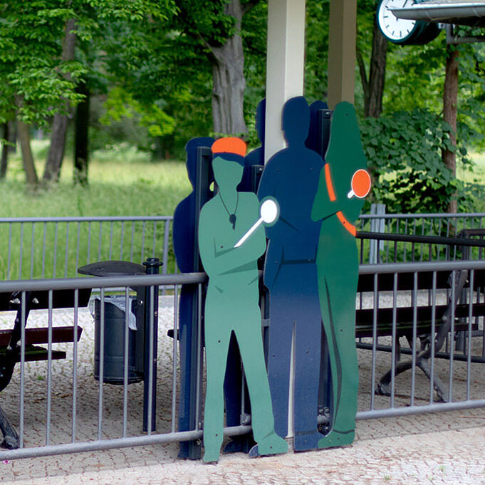 Lebensgroße Figuren von Menschen in Grün- und Blautönen stehen vor einem Zaun. Die Figuren tragen Schaffnerutensilien und verschränken die Arme.