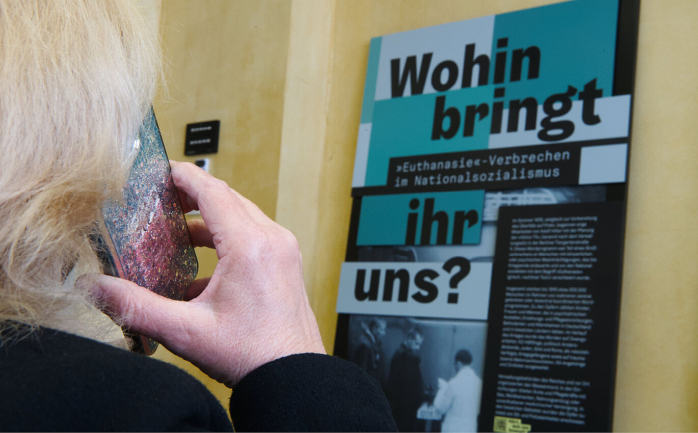 Eine Person steht vor einem der Rollups der Ausstellung und hört über ihr Handy den Audioguide.