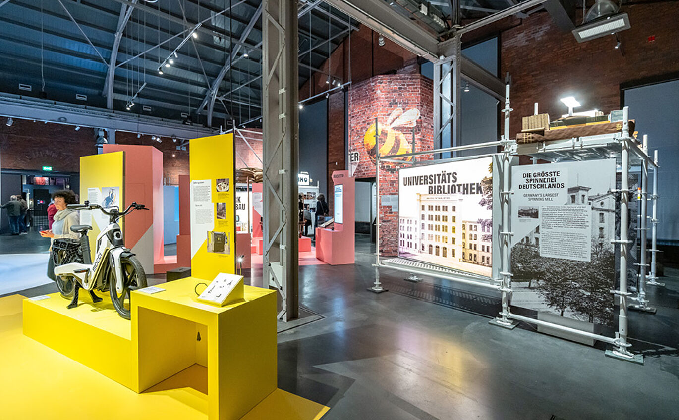 Realbild der Sonderausstellung. Vorne links ist ein gelb gehaltener Bereich mit Tischen zu erkennen, auf dem ein Fahrrad steht. Im Hintergrund sind Stahlstreben zu erkennen und mehrere Informationstafeln zu unterschiedlichen Themen.