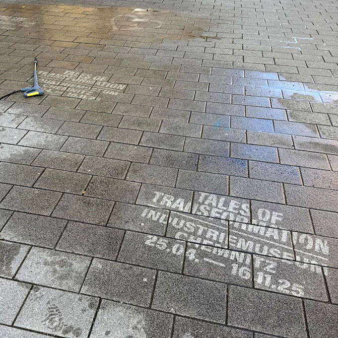 Realbild mehrerer Kreidestancels auf grauen Straßenplatten. Die Kreidestancels sind weiß und bewerben die Sonderausstellung Tales of Transformation.