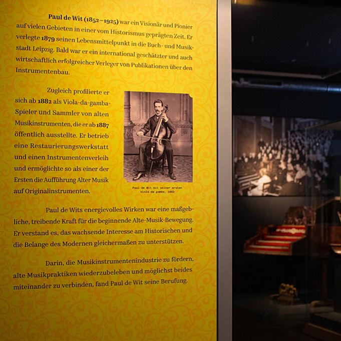 Informationstafel in der Leitfarbe ockergelb. Ein schwarz-weiß Foto von Paul de Wit wie er auf einem Instrument spielt ist von Text umgeben.