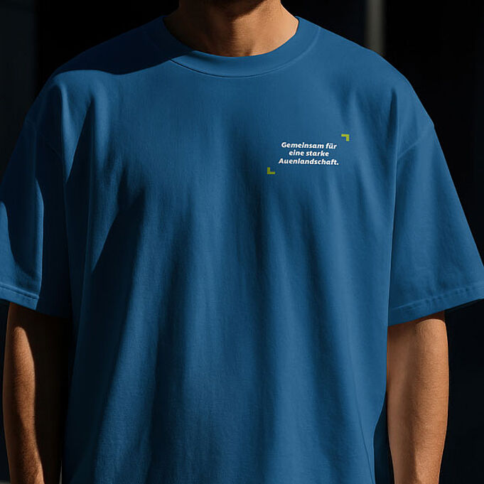 Mockup eines Tshirts an einer Person. Auf der Brust steht "Gemeinsam für eine starke Auenlandschaft.", eingerahmt von grünen Fokusecken. Das Tshirt ist blau.