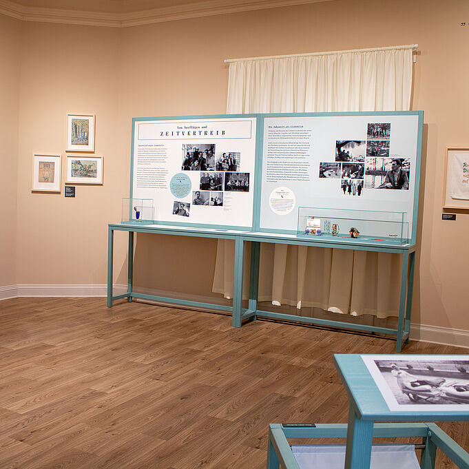 Ausstellungstische Hellblaue Infotafeln stehen an einer Wand. Auf einem kleinen Tisch der an den Tafeln dran ist, stehen Schaukästen mit Gegenständen.