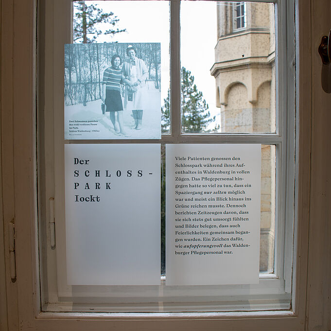 Fensterfolien Auf der inneren Scheibe eines alten, zweifach verglasten Fensters sind drei aufgeklebte Folien zu erkennen. Die oberste zeigt eine schwarz-weiß Fotografie einer Frau, die neben einer Krankenschwester steht und ihre Hand hält. Auf der Folie darunter steht "Der Schlosspark lockt". Auf der Folie unten rechts stehen Informationen zum Schlosspark und seiner damaligen Verwendung.