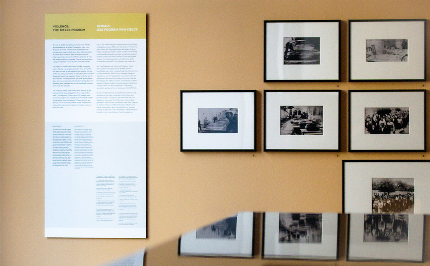 Vor einer orangenen Wand hängt links eine Informationstafel zu "Gewalt - Das Kielce Progrom". Rechts daneben hängen sechs eingerahmte schwarz-weiß Fotografien.