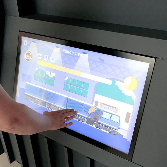 Ein Bildschirm ist in eine schwarz-graue Vorrichtung eingelassen, eine Hand schwebt darüber. Auf dem Bildschirm ist das Lokputzspiel zu erkennen.