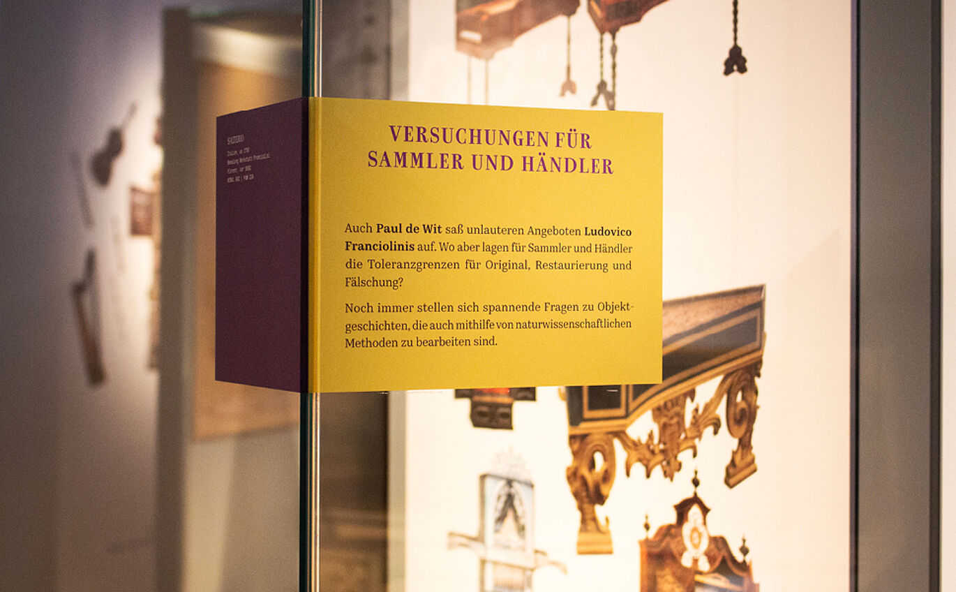 Nahaufnahme einer der Fensterfolien auf den Schaukästen innerhalb der Ausstellung. Die Folie ist über eine Ecke geklebt, die rechte Seite ist lila, die linke ockergelb. Die Überschrift lautet "Versuchungen für Sammler und Händler".