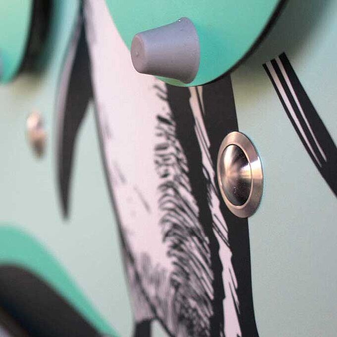 Nahaufnahme des Knopfes der Hörstation. Er ist rund und gold. Darüber ist ein Teil einer aufklappbaren Tafel zu sehen.