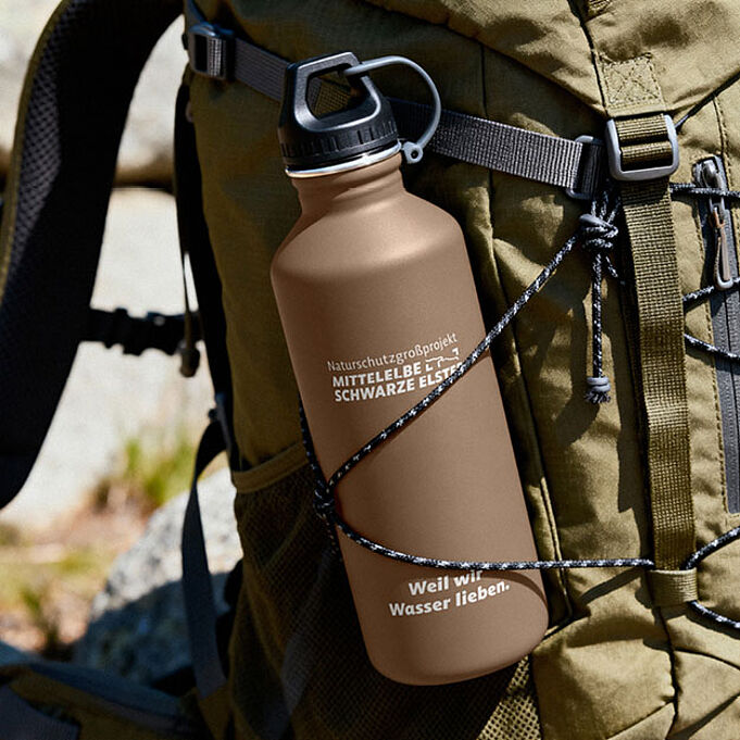 Mockup einer braunen Trinkflasche, die an der Seite eines Rucksacks befestigt ist. Auf der Flasche ist oben das Logo des Naturschutzgroßprojekt Mittelelbe - Schwarze Elster zu erkennen, darunter steht "Weil wir Wasser lieben.".