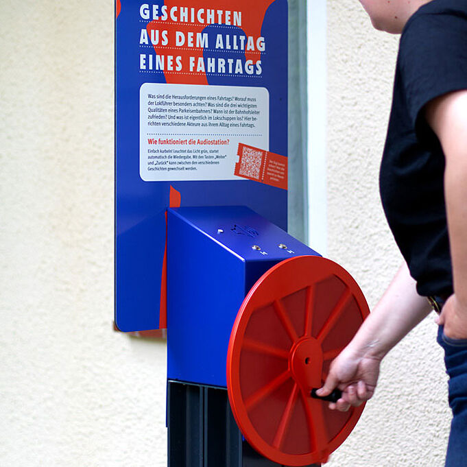 Die interaktive Vorrichtung "Geschichten aus dem Alltag eines Fahrtags" ist erneut zu sehen. Eine Frau dreht an dem roten Rad.