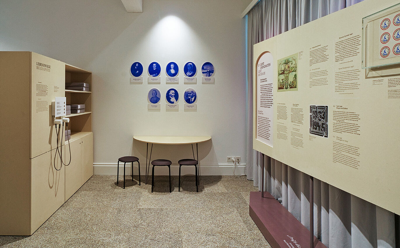 Rechts ist eine Informationstafel zu erkennen. An der Wand daneben sind mehrere blaue Portraits von Menschen, die Bilder sind in ovalen Rahmen. Darunter steht ein Tisch mit Stühlen. Links ist eine weitere Vermittlungsstation mit Hörelementen zu erkennen.