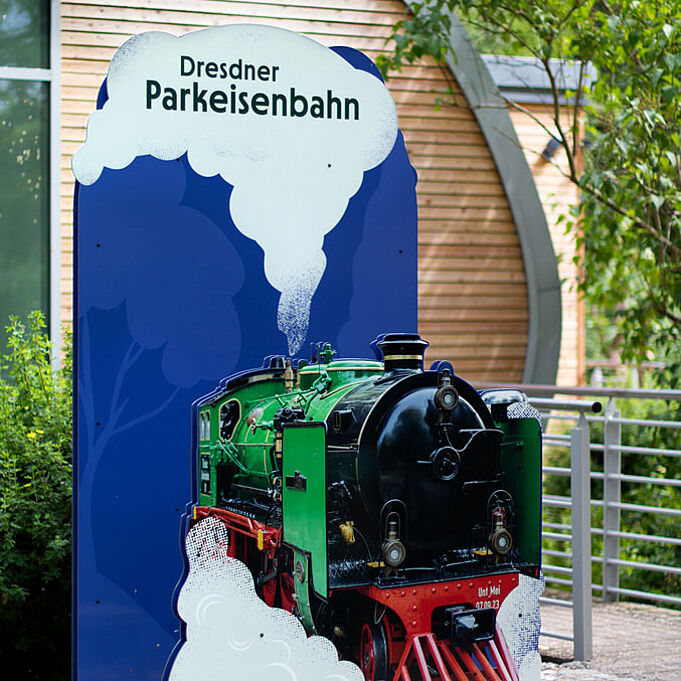 Ein blauer Aufsteller ist zu sehen. Im Vordergrund ist eine grün-rot-schwarte Dampeisenbahn zu sehen. Um die Bahn herum und über ihr ist gezeichneter weißer Dampf zu erkennen. Oben in der Dampfwolke steht "Dresdner Parkeisenbahn".