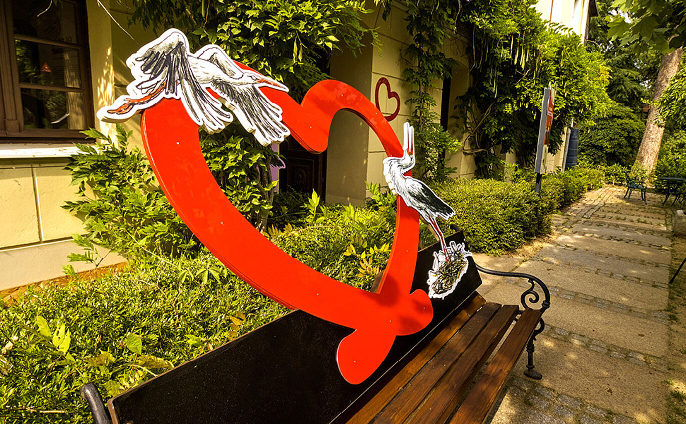 Eine Bank steht vor einer Hecke. Auf der Lehne ist ein großes rotes Herz zu erkennen, auf der rechten Seite ist die Illustration eines stehenden Storches abgebildet, von links fliegt ein Storch als Teil des Herzes heran.