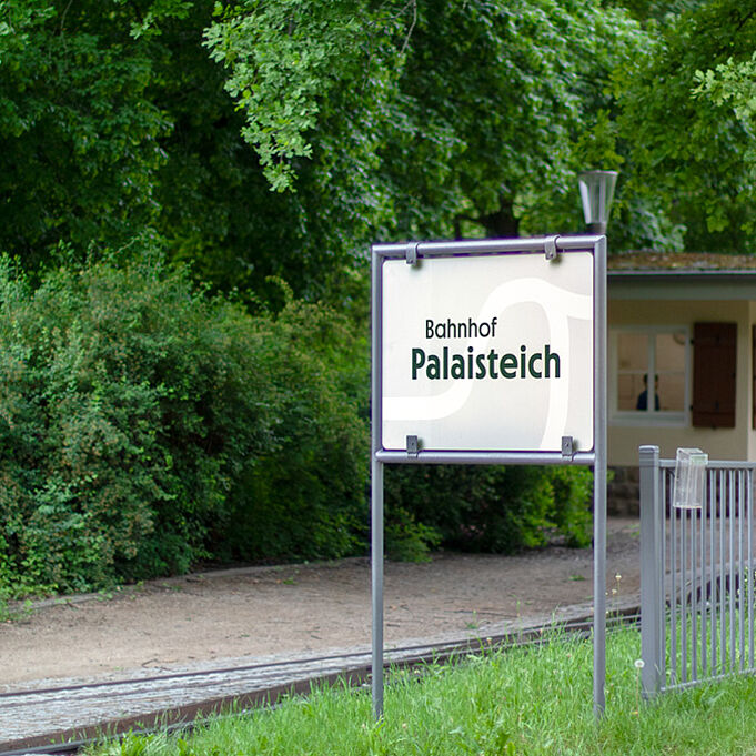Das Bild zeigt ein Schild mit der Aufschrift "Bahnhof Palaisteich". Im Hintergrund sind Bahnschienen und grüne Bäume zu erkennen.