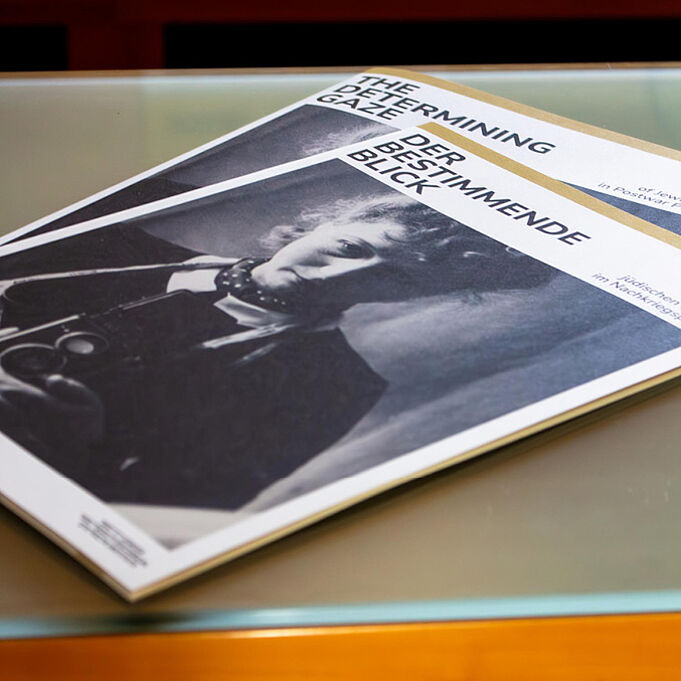 Zwei Flyer liegen auf einem Tisch. Sie leiten durch die Ausstellung. Auf dem Titelblatt ist erneut die junge Frau zu sehen. Der untere Flyer ist auf englisch, der obere auf deutsch.