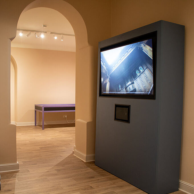 Filmmaterial Auf der linken Seite eines Raumes an der Wand steht ein großer grauer Vorsprung in den ein Bildschirm integriert ist. Auf dem Bildschirm sind Aufnahmen des Schloss Waldenburg zu erkennen. Im Hintergrund sind blaue Informationsplakate an der Wand.