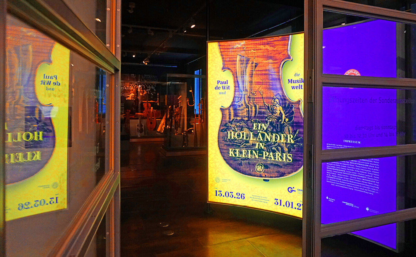 Blick durch den Eingang der Ausstellung. An den Seiten sind die geöffneten Glastüren zu sehen. Dahinter leuchtet die erste Lichttafel mit dem Key Visual und dem Namen der Ausstellung.