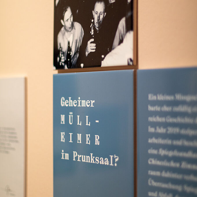 Fliesen Mehrere Fliesen an der Wand, bedruckt mit jeweils Teilen von Fotos lassen zwei Bilder nebeneinander entstehen. Auf dem linken Bild sind sieben Männer zu erkennen, die eng aneinander vor einem Weihnachtsbaum posieren und glücklich aussehen. Das rechte Bild zeigt fünf Männer, die scheinbar mehr oder weniger spontan fotografiert wurden. Vier schauen in die Kamera, einer von ihnen lächelt. Die Stimmung ist gut. Beide Bilder sind schwarz-weiß.
