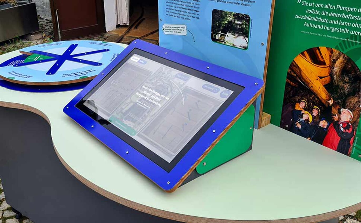 Nahaufnahme der Medienstation. Ein Monitor ist auf einem der wellenförmigen Tischen leicht schräg angebracht. Auf dem Bildschirm ist das von ungestalt entwickelte Spiel zu sehen.