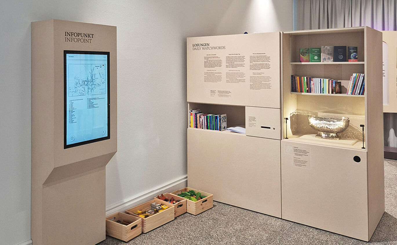 Foto des Infopunktes. Neben der Medienstation auf dem Boden stehen mehrere Kisten mit Spielzeug. Rechts daneben eine Vermittlungsstation zu dem Thema "Losungen".