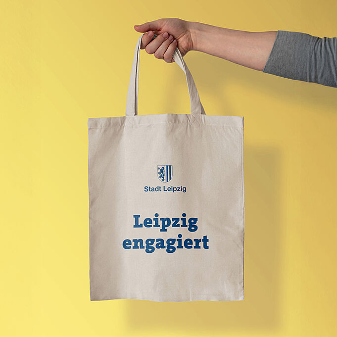 Ein Arm hält einen bedruckten Jutebeutel vor einer gelben Wand in die Kamera