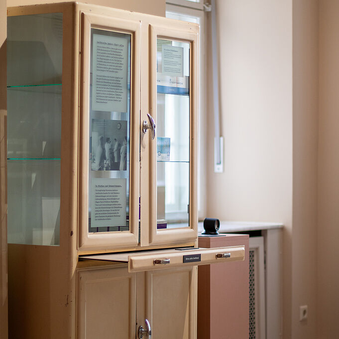 Originale Krankenhaus Ausstattung Ein alter Medizinschrank beinhaltet nachgebaute Verpackungen von alten Medikamenten. An den Scheiben ist oben eine Folie mit einem schwarz-weiß Bild von mehreren Ärzten zu erkennen, darunter eine Folie mit Fließtext und der Überschrift "Zu Risiken und Nebenwirkungen".