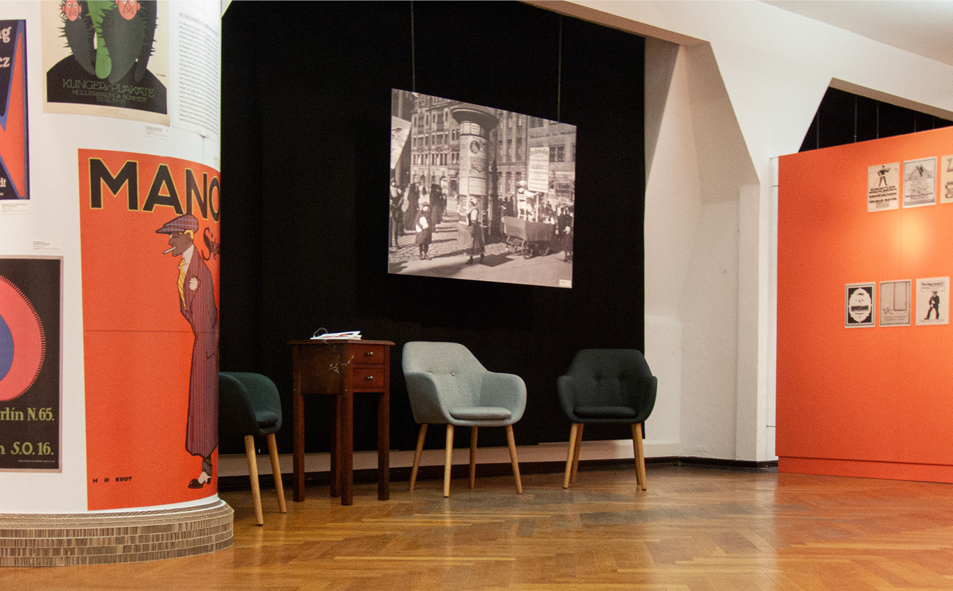 Next to an advertising column with typography poster are exhibits in the form of various pieces of furniture 