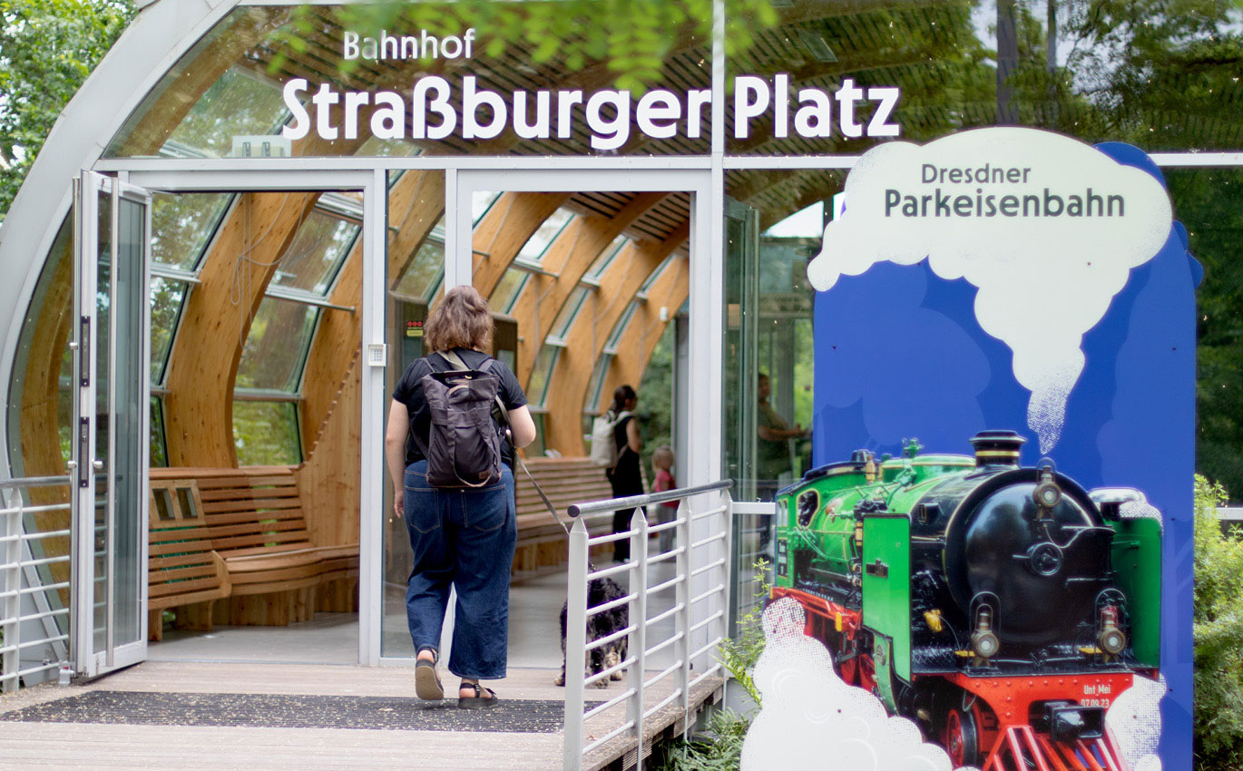 Eine Frau ist von hinten zu sehen. Sie läuft in ein Gebäude mit gläserner Front. Oben drüber steht "Bahnhof Straßburger Platz". Rechts im Bild ist ein blauer Aufsteller mit einer grünen Dampfeisenbahn darauf. Auf dem aufgezeichneten weißen Rauch steht "Dresdner Parkeisenbahn".