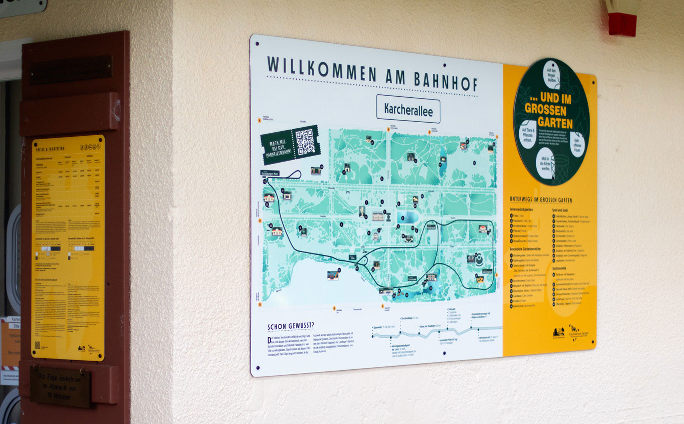 Eine große Informationstafel hängt an der Außenseite eines Gebäudes. Die Überschrift lautet "Willkommen am Bahnhof". Darunter steht klein "Karcherallee". Ein gezeichnetes Abbild der Parkanlage ist zu erkennen, in das in schwarz die Fahrlinie der Parkeisenbahnen eingezeichnet ist. Rechts in einem großen schwarten Kreis steht "...und im grossen Garten".