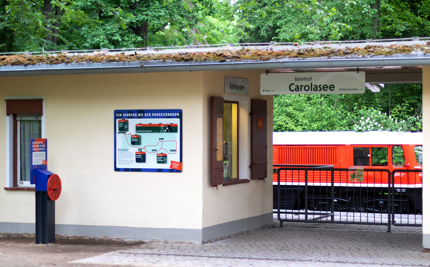 Ein weißes Haus steht zur Hälfte links im Bild. Daneben ist ein Zaun zu erkennen. Ein Vordach geht über den Zaun. Im Hintergrund steht eine rote Eisenbahn. Auf der Innenseite des Hauses ist ein Fenster zu erkennen, über dem ein Schild mit der Aufschrift "Fahrkarten" hängt. Neben dem Fenster sind hölzerne Fensterläden. Unter dem Dach hängt ein Schild mit der Aufschrift "Bahnhof Carolasee". Links an der Hausaußenseite ist eine Informationstafel angebracht mit der Überschrift "Ein Fahrtag bei der Parkeisenbahn". 