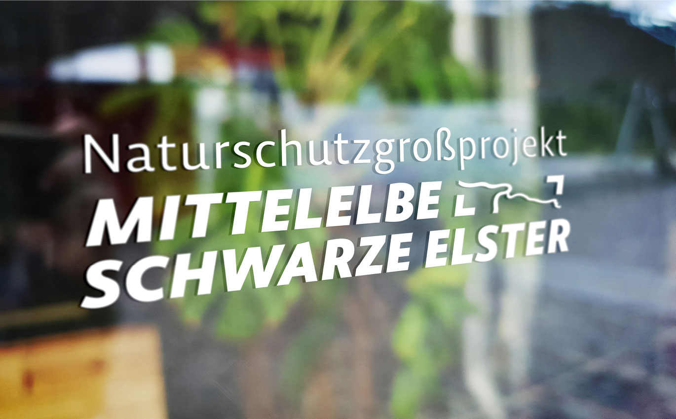 Das Logo des Naturschutzgroßprojekt Mittelelbe-Schwarze Elster ist in weiß auf eine Glasscheibe gedruckt.