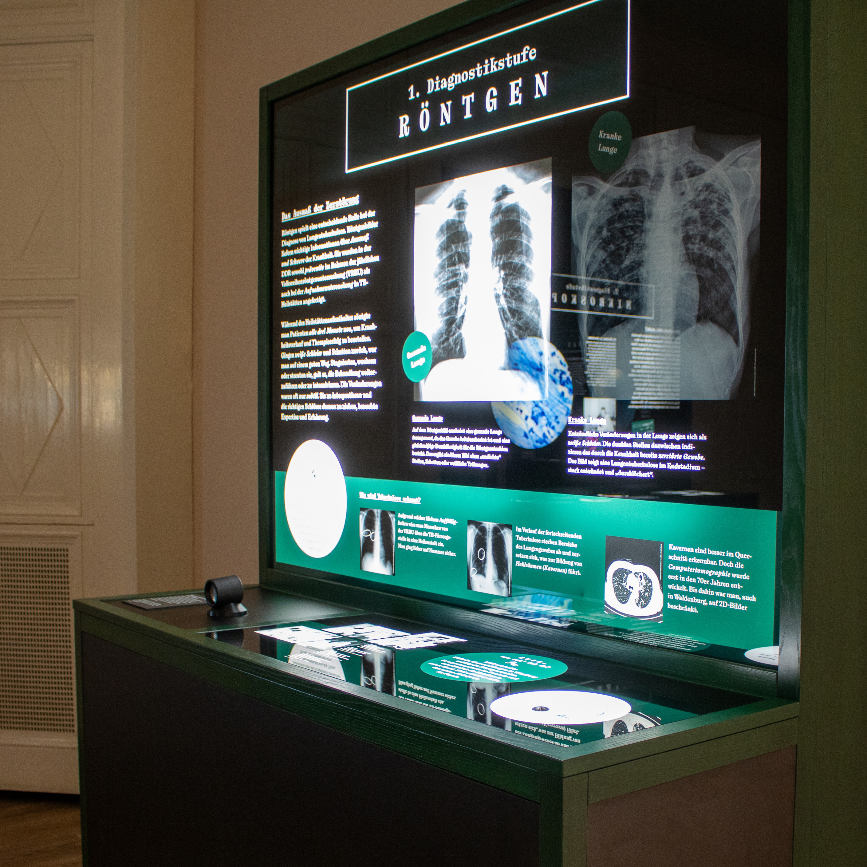 Leuchttafeln Auf einer dunklen Informationstafel, die von hinten beleuchtet wird, steht die Überschrift "3. Diagnostikstufe Bakterienkultur". Darunter sind kleinere Informationstexte zu erkennen. In der Mitte der Tafel ist ein großes, rundes, beleuchtetes Bild einer Bakterienkultur.