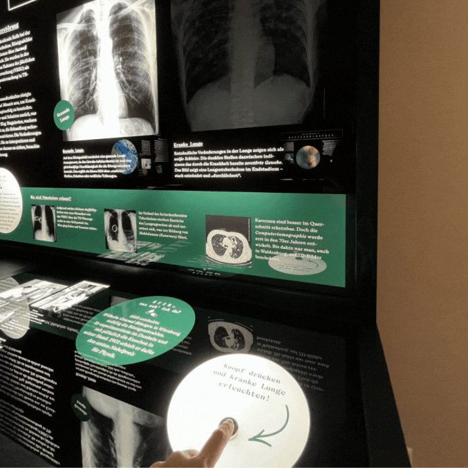 Entdecken Ein GIF bestehend aus drei Bildern. Auf der Bildabfolge ist zu erkennen, wie ein Finger auf einen Knopf drückt. Daraufhin leuchtet das Bild einer Lunge auf. Beides befindet sich auf einer dunklen Informationstafel die von hinten beleuchtet wird.