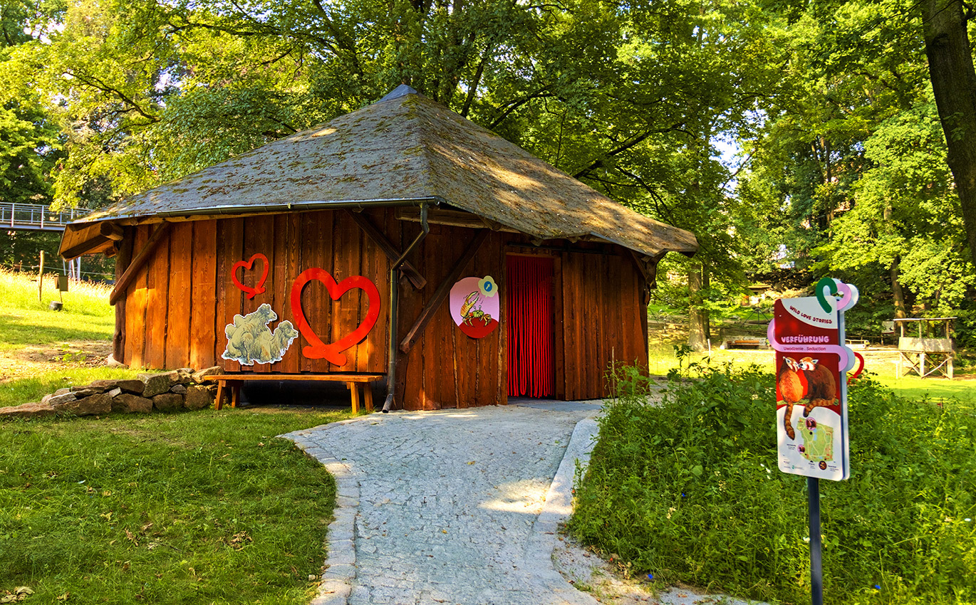 Foto einer der Ausstellungsgebäude von außen. Es handelt sich um ein kleines Holzhaus. Links an der Außenwand ist eine Bank, über der zwei Herzen und zwei Kamele aus Holz angebracht sind.
