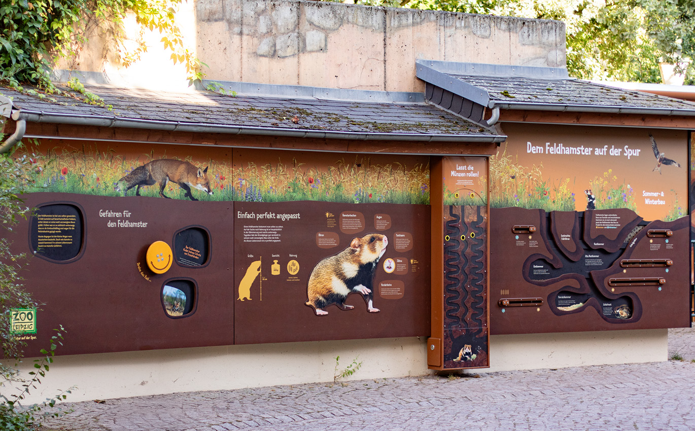 Realaufnahme der Hamsterwand im Weitwinkel. Es ist zu erkennen, dass die Informationen an einer Hauswand angebracht sind. Die Überschrift lautet "Dem Feldhamster auf der Spur".