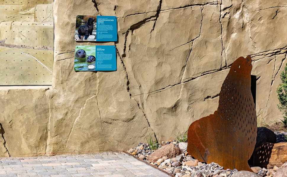 Foto einer Stahlsilhouette einer Robbe neben Gehegeschildern.